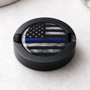 May include: A round black ashtray featuring a circular insert with a distressed American flag design. The flag is rendered in shades of black, white, and gray, with a single horizontal blue stripe running across the center of the stripes.