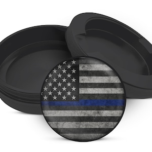May include: A black container with a lid and a round insert featuring a distressed American flag design with a blue stripe. The flag design includes white stars and stripes, with a blue stripe across the center.