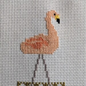 Pode incluir: Ponto cruz de um flamingo com corpo pêssego, bico amarelo e ponta preta. O flamingo está em pé sobre pernas cinzentas acima de uma linha costurada verde e castanha, possivelmente representando água. O fundo é um tecido branco de ponto cruz.