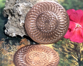 May include: Two copper coasters with a circular, intricate, mandala-like design. The coasters are on a background of green and brown foliage.