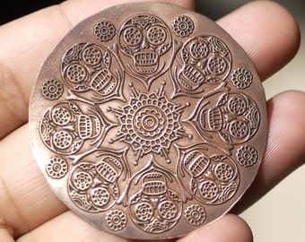 May include: A copper-colored metal disc with a circular design featuring eight stylized skulls with intricate details. The skulls are arranged in a circular pattern around a central mandala-like design.