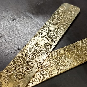 May include: Two gold-colored metal bookmarks with intricate paisley and floral patterns. The bookmarks are long and narrow, with a slightly rounded edge.