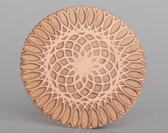 May include: A round, copper-colored metal coaster with a geometric design. The design features a series of concentric circles with a repeating pattern of overlapping ovals.