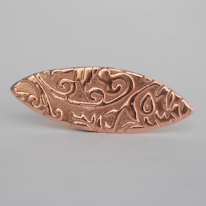 Puede incluir: Un colgante de forma ovalada de color cobre con un diseño detallado, en espiral y en relieve.