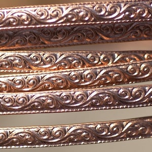 May include: Close-up of several copper-coloured metal bands. Each band features an ornate, repeating pattern of swirling floral designs. The bands are slightly curved, suggesting they may be part of a larger decorative item or jewellery.