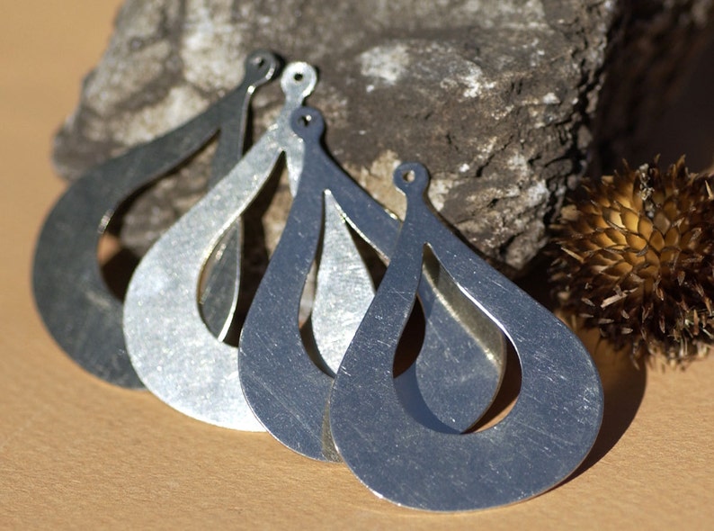May include: Four silver teardrop-shaped earrings with a cutout design. The earrings are arranged in a row, with the largest earring at the bottom and the smallest earring at the top.