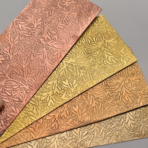 May include: Four rectangular metal plates with embossed leaf patterns in shades of copper, gold, and bronze. The plates are arranged in a fan shape, showcasing the detailed floral design. The plates have a textured surface.