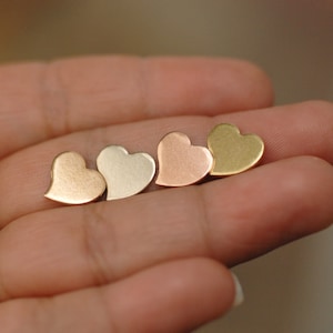 May include: Four heart-shaped stud earrings in varying metallic finishes: gold, silver, rose gold, and brass. The earrings are small and delicate, showcasing a simple, romantic design. The earrings are displayed on a light background.