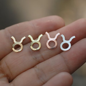 May include: Four small metal Taurus symbol charms in gold, brass, rose gold, and silver. Each charm features a circular body with curved horns, representing the astrological sign. The charms are displayed against a blurred background.