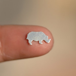 May include: A small, silver-colored rhinoceros silhouette charm, shown on a finger.