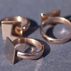 Puede incluir: Tres anillos de color dorado con tapas cuadradas. Los anillos están apilados sobre una superficie azul.