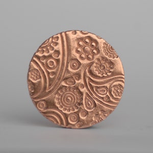 Puede incluir: Un colgante redondo de color cobre con un diseño paisley intrincado en relieve. El diseño presenta remolinos, círculos y motivos florales.