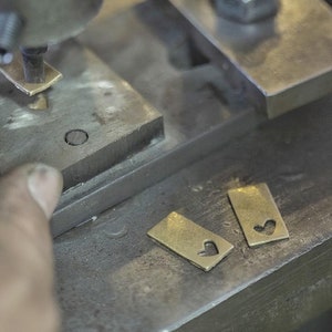 以下が含まれることがあります： ハート型の切り抜きが施された金色の金属製のタグが2つ、灰色の金属製の表面に置かれています。背景には金属製のプレスが見えます。
