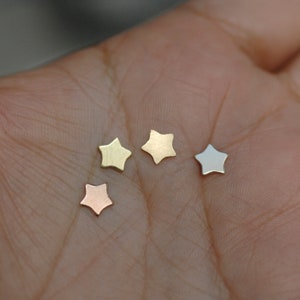 May include: Four small star-shaped charms in different colors: gold, silver, rose gold, and copper. The charms are displayed on a hand.