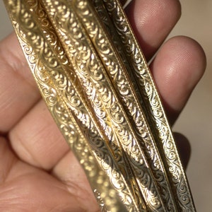 May include: Close-up of four gold-colored metal bands with intricate, swirling patterns. The bands are stacked on top of each other and are held in a hand.