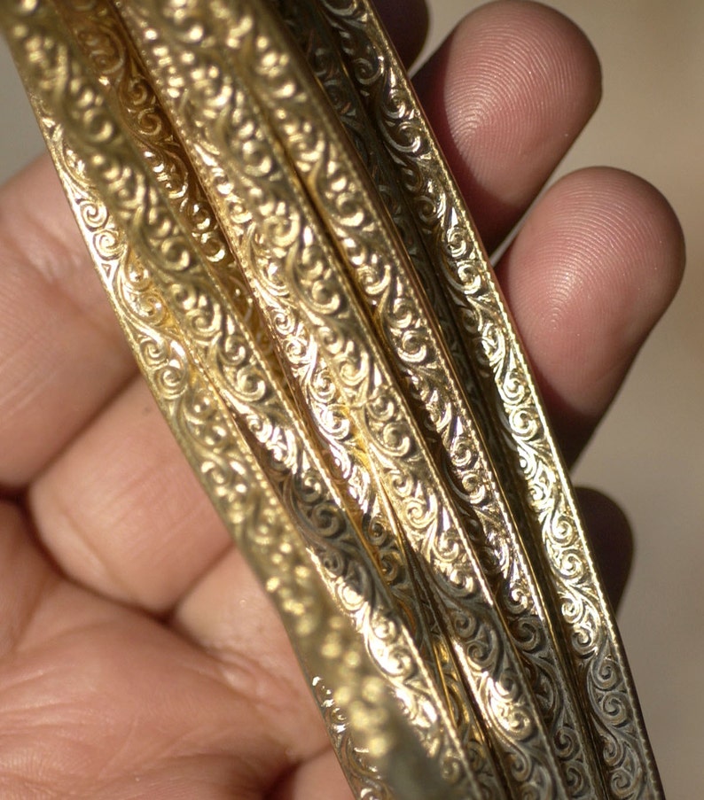 May include: Close-up of four gold-colored metal bands with intricate, swirling patterns. The bands are stacked on top of each other and are held in a hand.