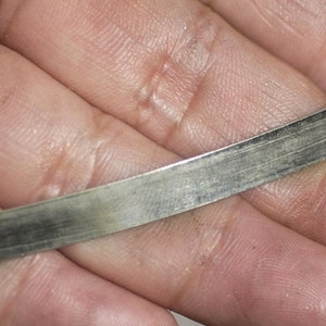 May include: A close-up of a thin, metallic strip with a brushed texture. The strip appears to be silver or gray in color, with visible lines running along its length. The object is held in a hand, suggesting a tool or component.