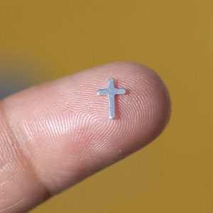 May include: A small silver cross charm, held between a finger and thumb.