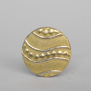 Op de afbeelding: Een gouden knoop met een golvend ontwerp en een rij kleine cirkels rond de rand.