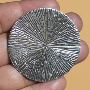 May include: A silver circular pendant with a textured, radial pattern. The pendant has a smooth, polished finish.