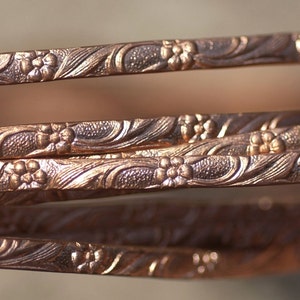 Op de afbeelding: Close-up van verschillende koperkleurige armbanden. Elke armband heeft een gedetailleerd bloemmotief, met kleine bloemen en wervelende bladeren. De armbanden zijn gestapeld, waardoor een gelaagd effect ontstaat. De warme tinten en het ingewikkelde ontwerp suggereren een vintage of handgemaakte esthetiek.