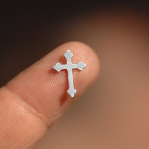 May include: A small, silver cross with a simple design is held on a finger.
