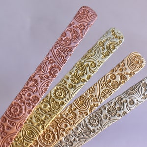 May include: Four metal strips with a paisley pattern embossed on the surface. The strips are in shades of copper, gold, silver, and white.
