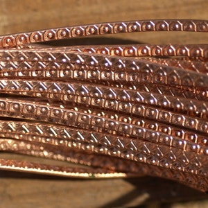 May include: Close-up of a coil of copper wire with a decorative, textured surface. The wire has a warm, reddish-brown color and a repeating pattern of small squares and circles.