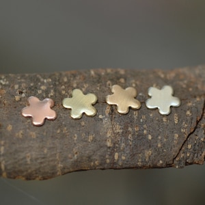 Könnte beinhalten: Vier kleine, blumenförmige Charms aus Metall in verschiedenen Farben: Kupfer, Gold, Silber und Weißgold.