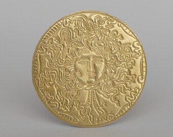 May include: A gold-colored round medallion with an embossed image of a woman's face with curly hair. The woman's face is surrounded by a decorative border.