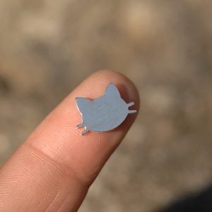 May include: A silver metal charm in the shape of a cat's face. The charm is small and has a simple design.