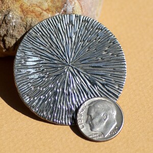 May include: A silver circular pendant with a textured, sunburst design. The pendant is about the size of a dime.