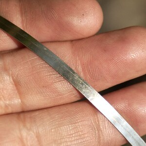 May include: A thin, metallic strip with a slightly reflective surface. The strip is held in a hand, with the texture of the skin visible in the background. The metal appears to be silver or a similar light color.