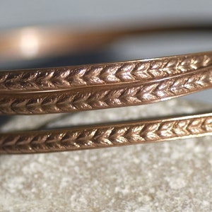 May include: Close-up of a copper-colored bracelet with a detailed leaf pattern. The bracelet is coiled and rests on a light gray surface. The intricate design and warm color suggest a handcrafted piece of jewelry.