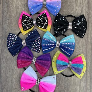Peut inclure: Collection d'accessoires pour cheveux en forme de nœuds colorés avec des strass. Les nœuds présentent diverses couleurs, dont le rose, le bleu, le violet et le noir, avec des effets pailletés et ombrés. Chaque nœud est fixé à un élastique noir.