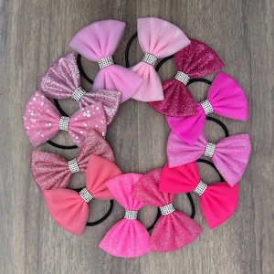 Può includere: Una collezione di elastici per capelli rosa disposti a cerchio. I fiocchi presentano varie tonalità di rosa, alcuni con dettagli glitterati e paillettes. Ogni fiocco è adornato da una fascia tempestata di strass. Lo sfondo è una superficie in legno.