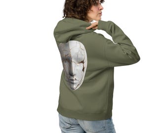 Peut inclure: Un sweat à capuche vert olive avec un motif de visage divisé sur le dos. Le visage est divisé en deux moitiés, grise et blanche, avec des fissures. Le sweat à capuche a une capuche et des manches longues. Le modèle porte un jean bleu.