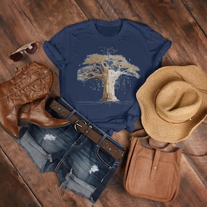 Op de afbeelding: Een marineblauw t-shirt met een boomafbeelding, gecombineerd met een spijkerbroek, een bruine riem, cowboylaarzen, een strohoed, een zonnebril en een bruine leren tas. De items zijn gerangschikt op een houten oppervlak.