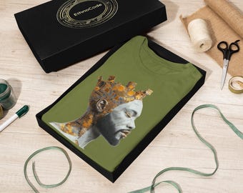 Può includere: T-shirt verde oliva con un ritratto di profilo di una persona con una corona. Il ritratto ha accenti oro e grigio. La maglietta è in una scatola nera con il logo EthnoCode sul coperchio. Altri oggetti includono un barattolo di vernice verde, un pennarello, forbici e nastro.
