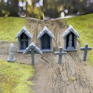 Puede incluir: Cinco estructuras de madera pequeñas de color gris, incluyendo tres pequeñas casas con puertas abiertas y dos cruces, están dispuestas en un camino de tierra con hierba verde a cada lado.