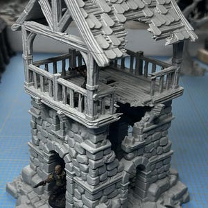 Può includere: Una miniatura grigia di una torre in rovina, stampata in 3D, con un tetto rotto e una piccola figura in piedi nell'ingresso. La torre è in pietra e ha un balcone in legno.