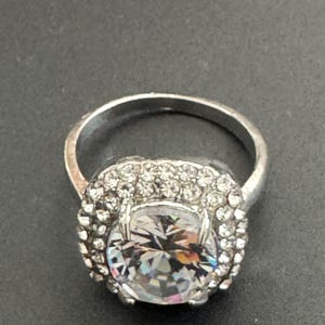 Op de afbeelding: Een zilverkleurige ring met een grote, heldere, gefacetteerde edelsteen in het midden, omringd door kleinere, heldere stenen. De ring heeft een klassiek, elegant ontwerp, geschikt voor diverse gelegenheden.