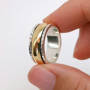 Könnte beinhalten: Ein zweifarbiges Ring mit einem goldfarbenen Mittelband und silbernen Außenbändern. Der Ring wird zwischen zwei Fingern vor einem weißen Hintergrund gehalten. Das goldfarbene Band hat eine polierte Oberfläche, während die silbernen Bänder ein strukturiertes Design aufweisen.