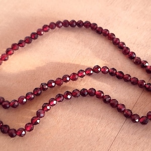 May include: A strand of dark red gemstone beads, likely garnet, strung on a thin red thread. The beads are faceted and have a shiny, polished finish.