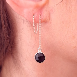 Op de afbeelding: Een zilveren kettingoorbel met een zwarte edelsteen die aan het uiteinde hangt. De oorbel wordt in het oor van een persoon gedragen.