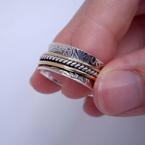 Könnte beinhalten: Ein silberner und goldener Spinner-Ring mit aufwendigen Details. Der Ring hat ein silbernes Band mit einem Muster und zwei goldene Bänder. Der Ring wird in einer Hand vor weißem Hintergrund gehalten.
