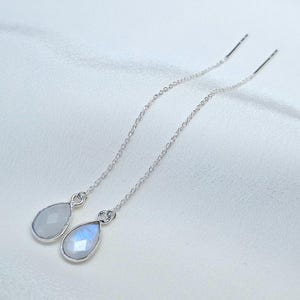 Könnte beinhalten: Silberne Ohrhänger mit tropfenförmigen Mondsteinanhängern. Ein Anhänger ist hell und undurchsichtig, der andere zeigt einen schimmernden, irisierenden Effekt. Die Ohrringe hängen vor einem weißen, seidigen Hintergrund.