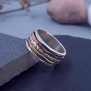 Op de afbeelding: Een zilveren ring met meerdere banden in verschillende kleuren en texturen. De ring heeft een band met kleine, heldere stenen, een koperkleurige band en een goudkleurige band. De ring staat tegen een donkere achtergrond.