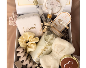 May include: A white gift box containing a personalized jewelry case with the name Emma, a gold bangle, a glass vial with dried flowers, a dropper bottle, two hair clips, a cream scrunchie, a candle labeled Emma, a headband, and a compact mirror.