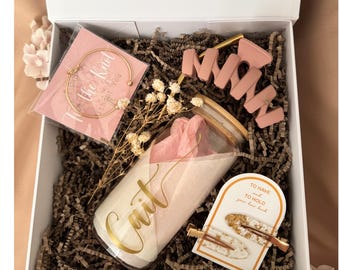 May include: A white gift box filled with brown paper crinkle filler containing a clear glass tumbler with a bamboo lid and the name Cait in gold script, a gold bangle on a card, a dusty pink hair claw, and two gold-flecked hair clips on a card.